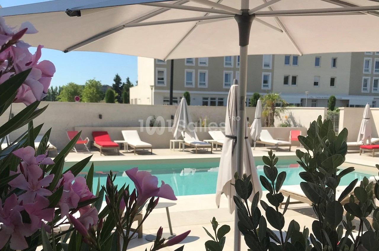 Piscine extérieure entourée de chaises longues et parasols, avec des fleurs au premier plan et un bâtiment en arrière-plan.