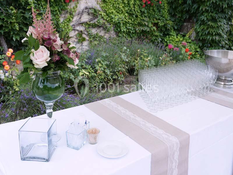 Table dressée en extérieur avec nappes blanches, verres empilés, vase fleuri et décor végétal en arrière-plan.