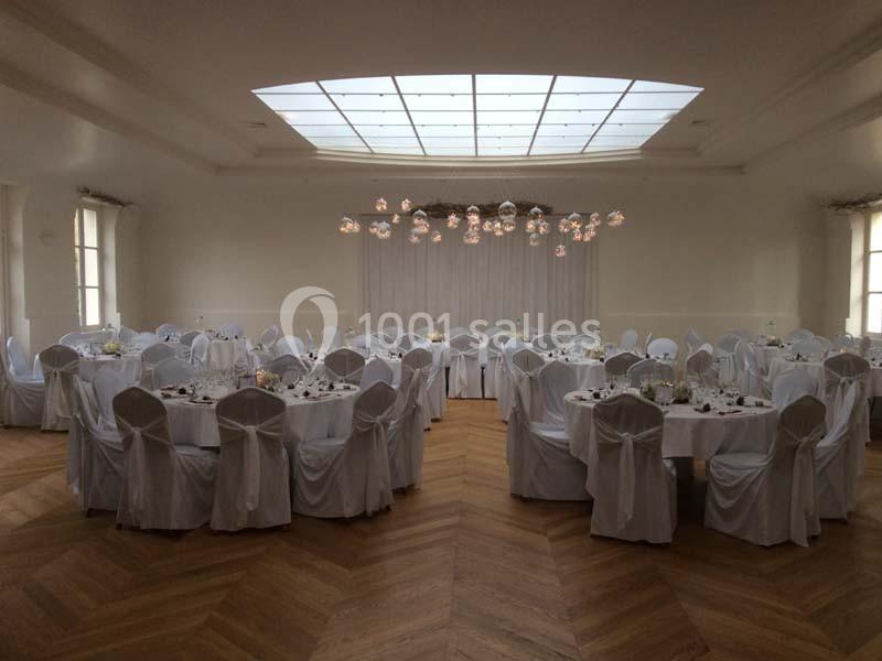 Salle de réception lumineuse avec tables rondes dressées et chaises couvertes de housses blanches.