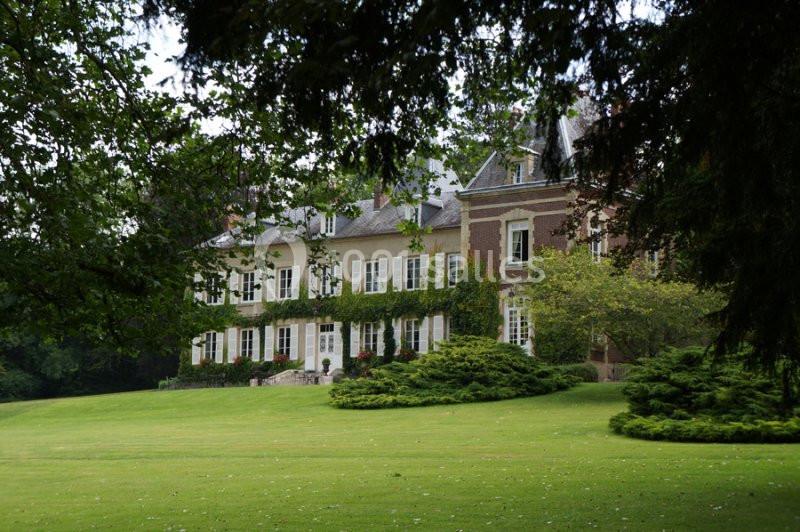 Grande maison en pierre et briques entourée d'un vaste jardin verdoyant avec pelouse et arbres.