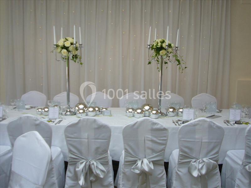 Table de réception élégamment dressée avec nappes blanches, chaises décorées et centres de table fleuris.