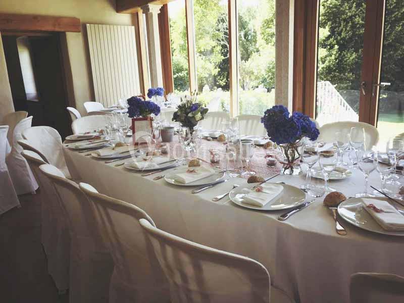 Table dressée pour un repas, avec nappes blanches, vaisselle élégante et bouquets de fleurs bleues, près de grandes fenêtres.