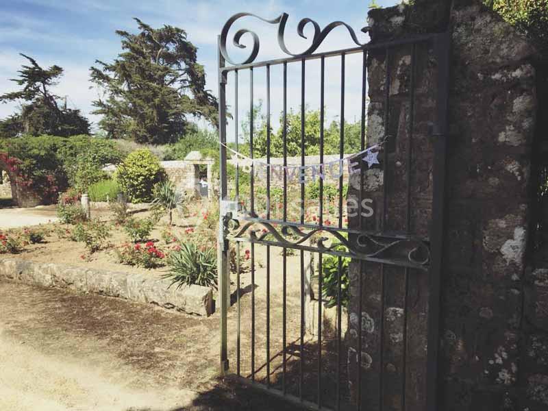 Portail en fer forgé ouvert, entouré de pierres, donnant sur un jardin fleuri par une journée ensoleillée.