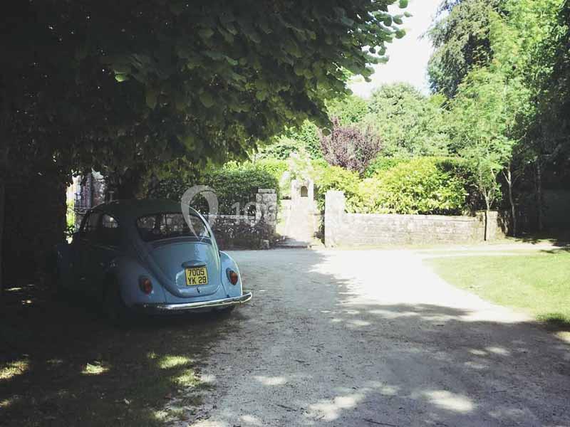 Une voiture ancienne bleue garée à l'ombre d'un arbre près d'une allée bordée de verdure et d'un mur en pierre.