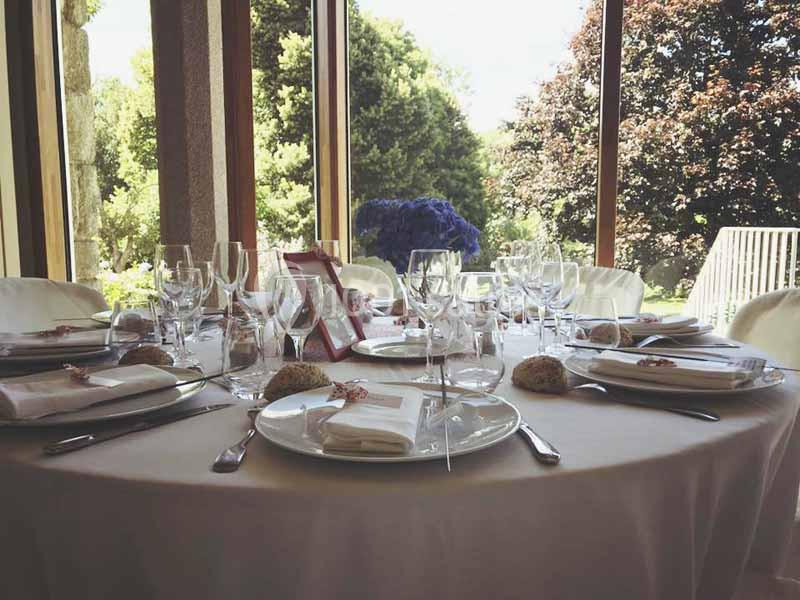 Table élégamment dressée avec nappes blanches, vaisselle raffinée et vue sur un jardin arboré à travers de grandes baies…