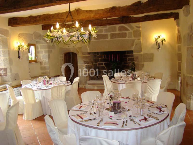 Salle de réception rustique avec poutres apparentes, tables rondes dressées et cheminée en pierre.