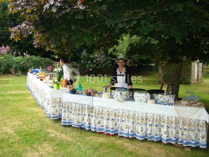 Table dressée en extérieur sous des arbres, avec nappes décorées, plats variés et une personne en tenue traditionnelle.