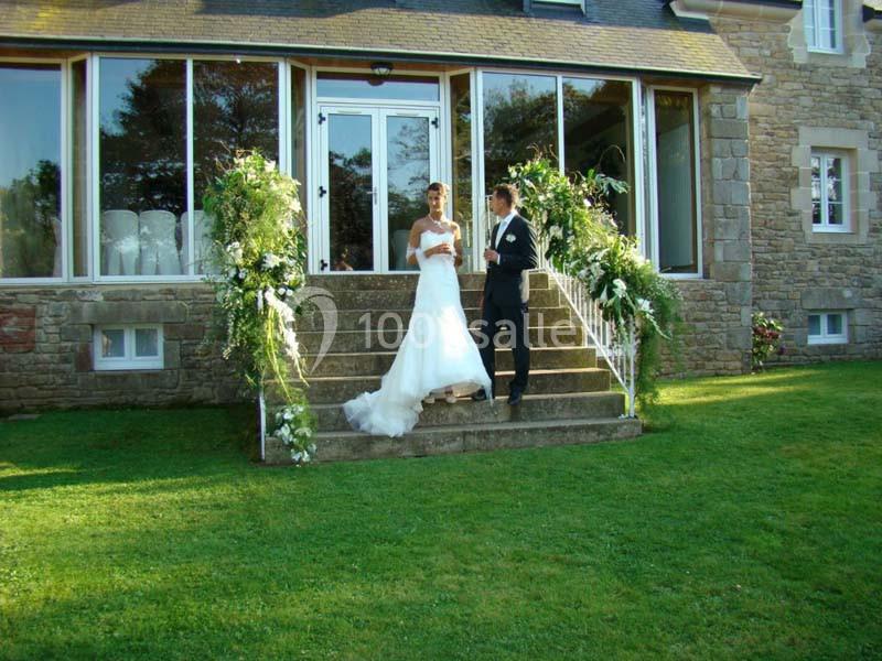 Un couple en tenue de mariage pose sur les marches fleuries devant une maison en pierre avec de grandes fenêtres.