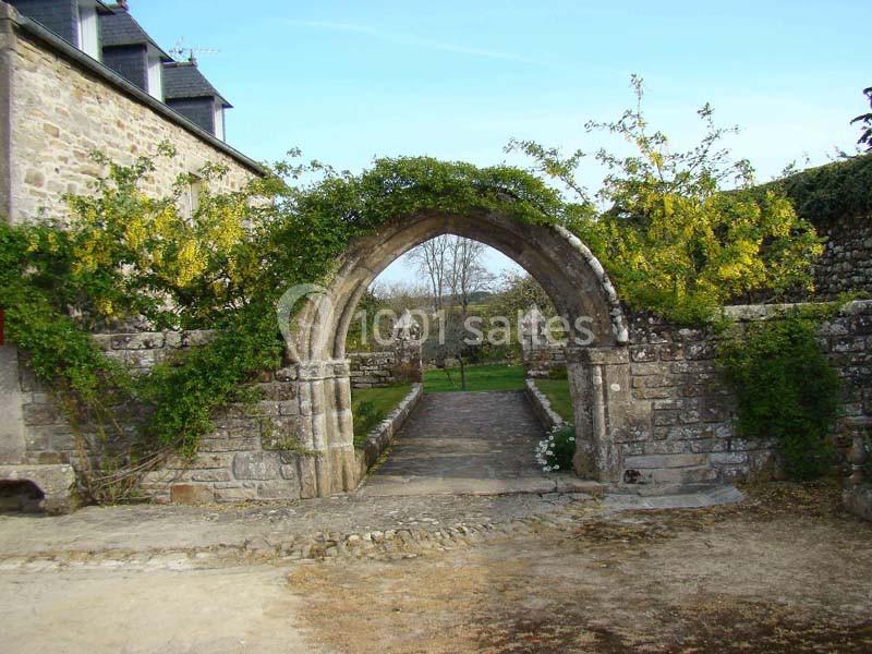 Arche en pierre ancienne entourée de végétation, donnant sur un chemin bordé de murs et un paysage champêtre.