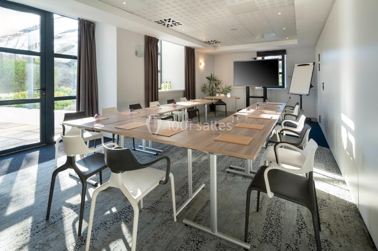 Salle de réunion lumineuse avec tables disposées en U, chaises modernes, écran, paperboard et grandes fenêtres.