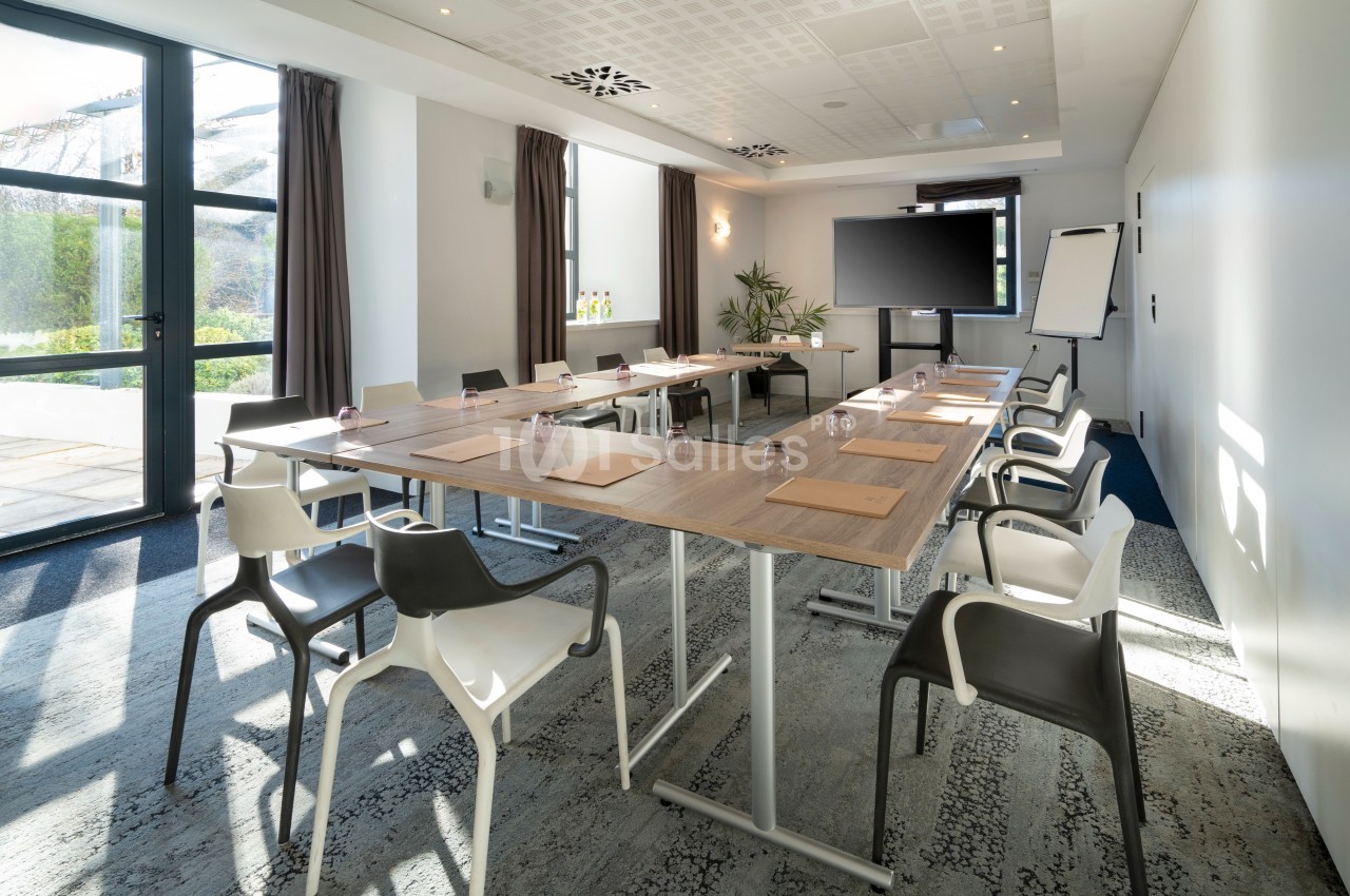 Salle de réunion lumineuse avec tables disposées en U, chaises modernes, écran, paperboard et grandes fenêtres.