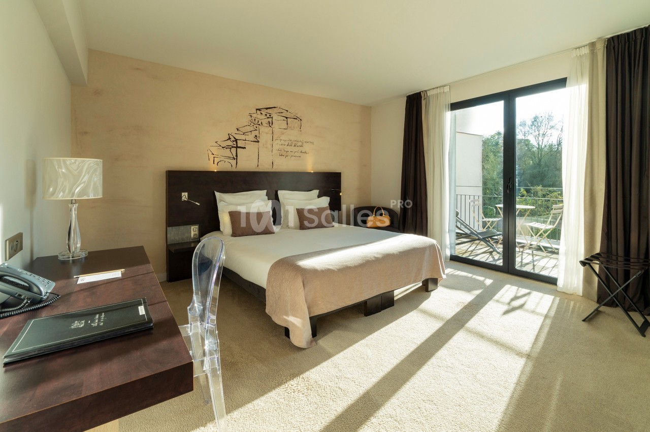 Chambre lumineuse avec lit double, bureau, chaise transparente et balcon donnant sur un espace extérieur verdoyant.