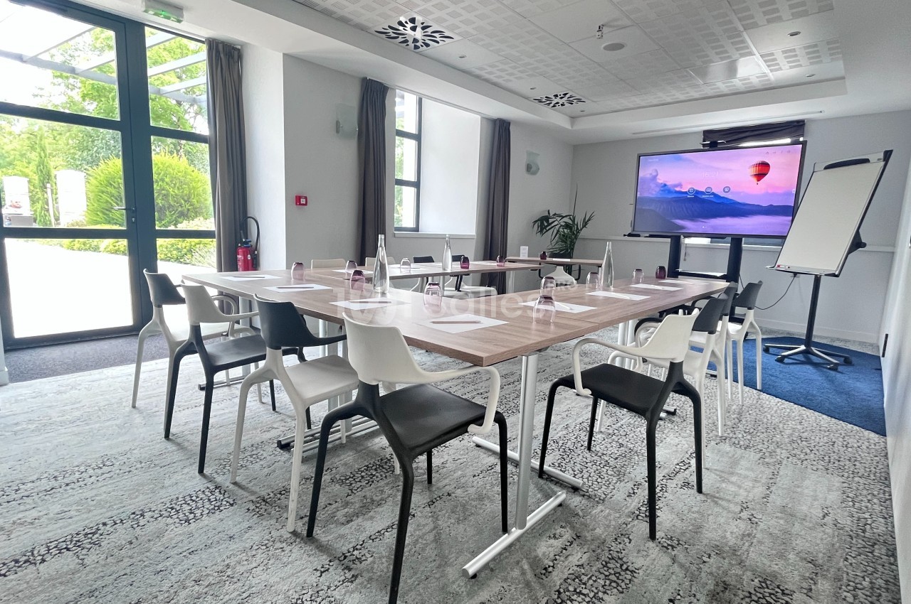 Salle de réunion lumineuse avec table rectangulaire, chaises, écran de présentation, tableau blanc et grandes fenêtres.