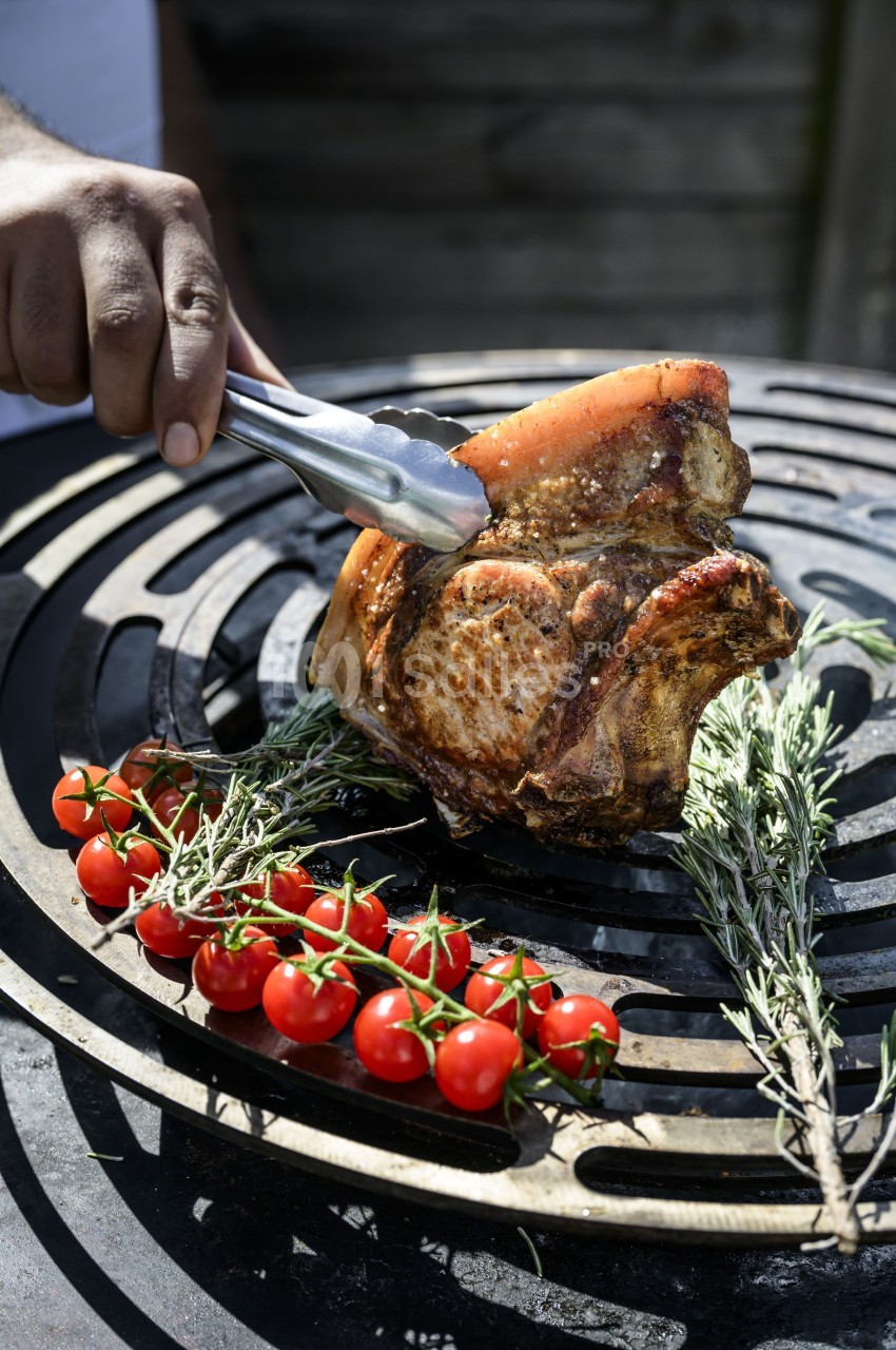 Viande grillée sur un barbecue avec des tomates cerises et des herbes fraîches, tenue par une pince en métal.