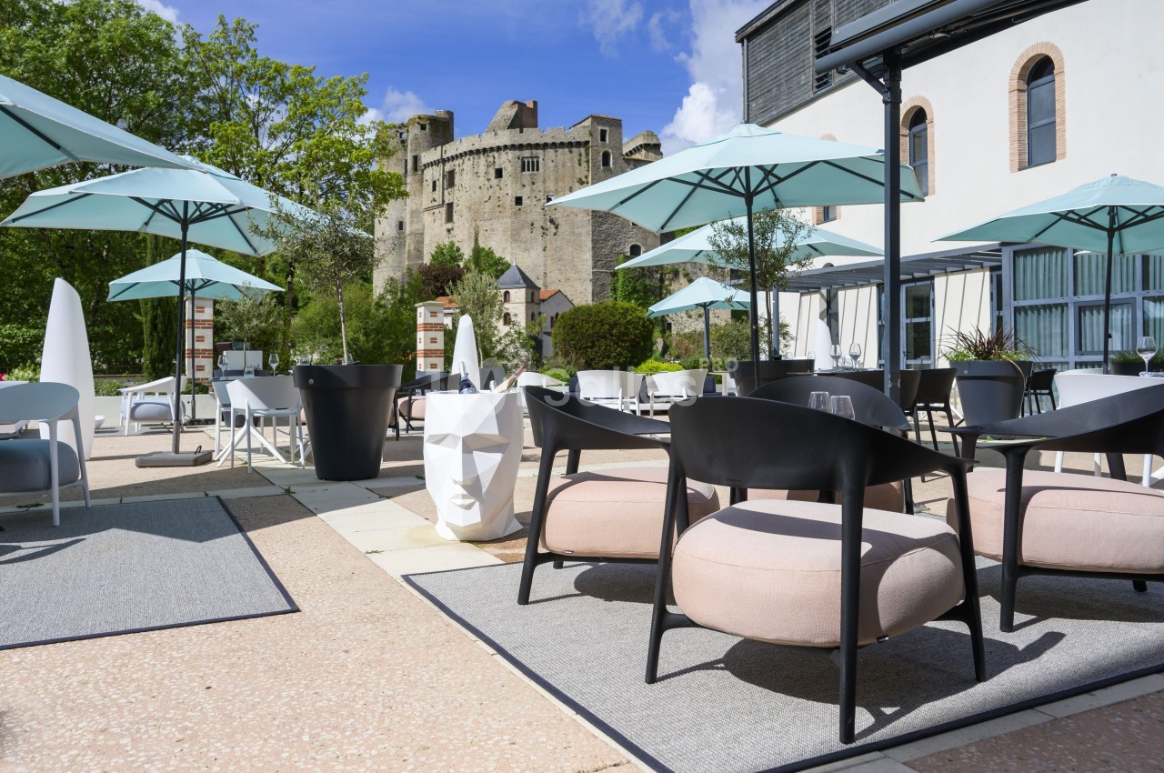 Terrasse aménagée avec mobilier moderne, parasols et vue sur un château médiéval sous un ciel ensoleillé.