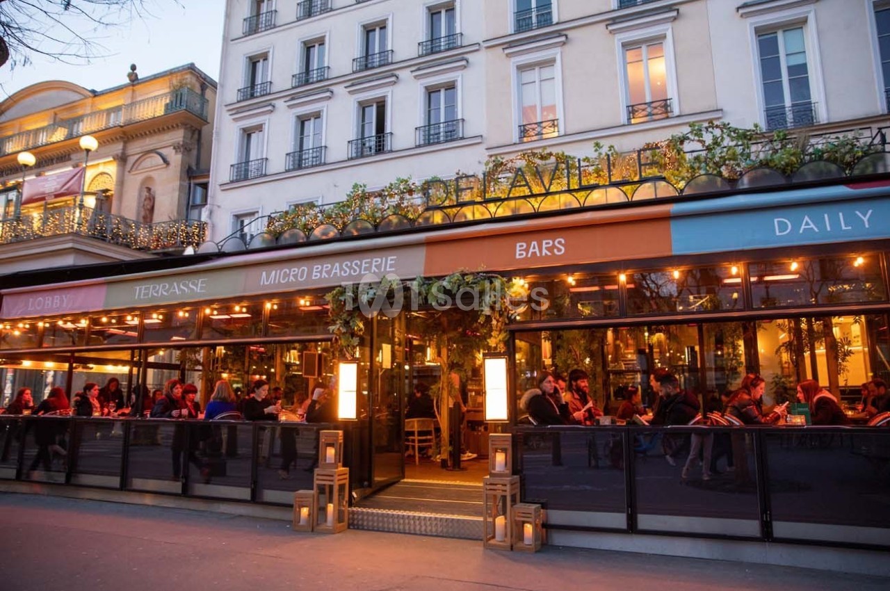 Façade d'un café-restaurant animé avec terrasse éclairée, situé au rez-de-chaussée d'un immeuble parisien.