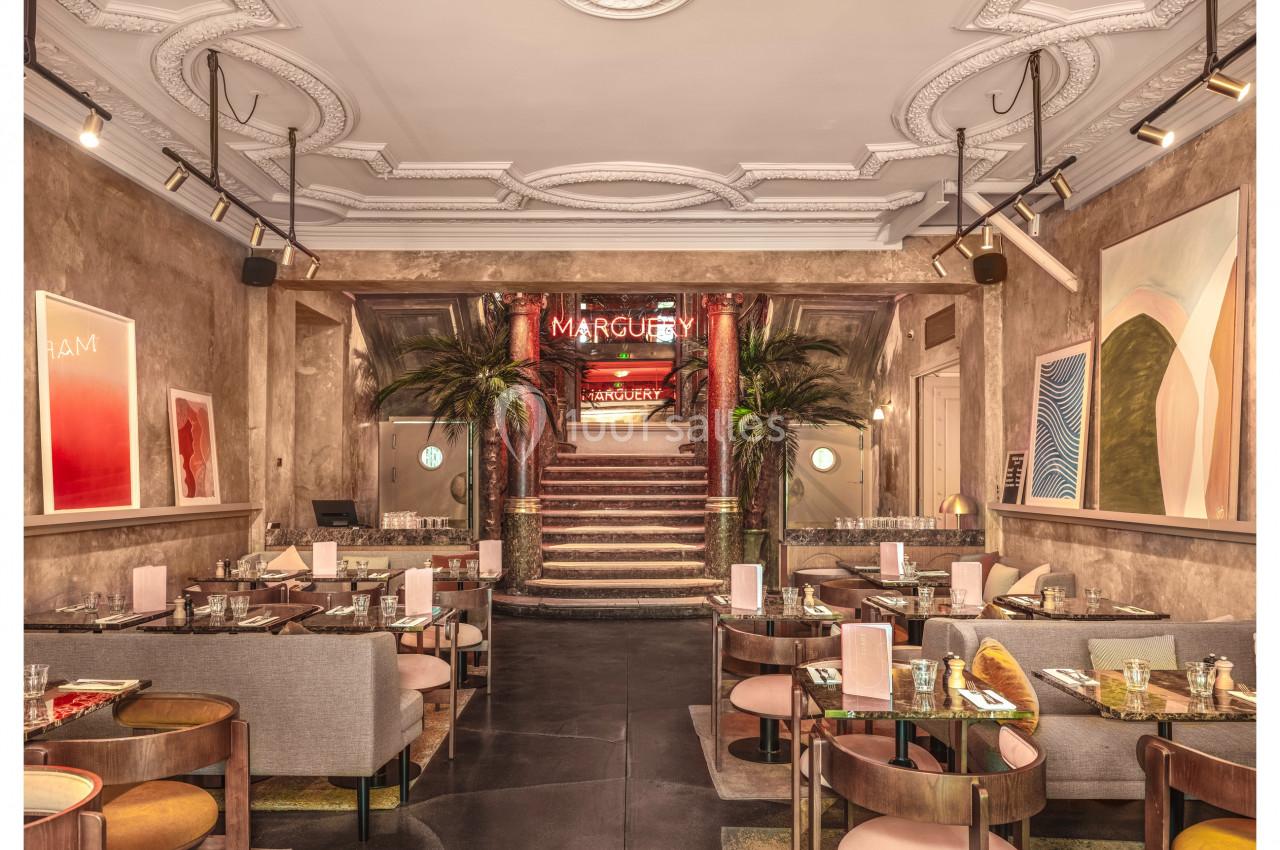 Salle de restaurant élégante avec des tables dressées, un escalier central, des plantes et des œuvres d'art aux murs.