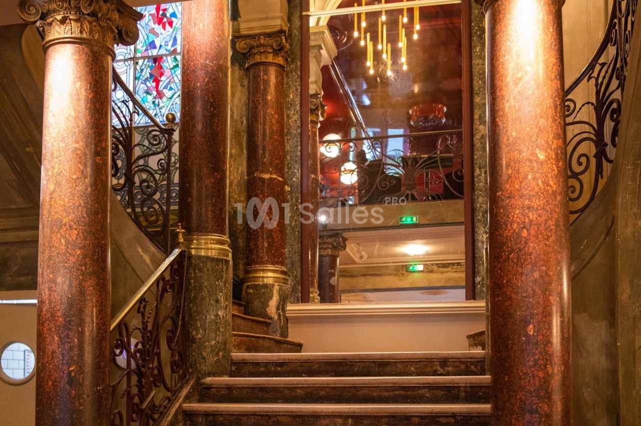 Escalier en marbre entouré de colonnes, avec une rambarde en fer forgé et un vitrail coloré en arrière-plan.