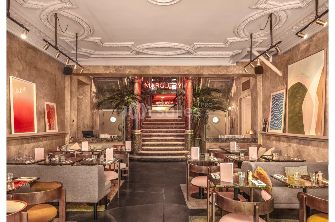 Salle de restaurant élégante avec tables dressées, décoration moderne et escalier central entouré de palmiers.