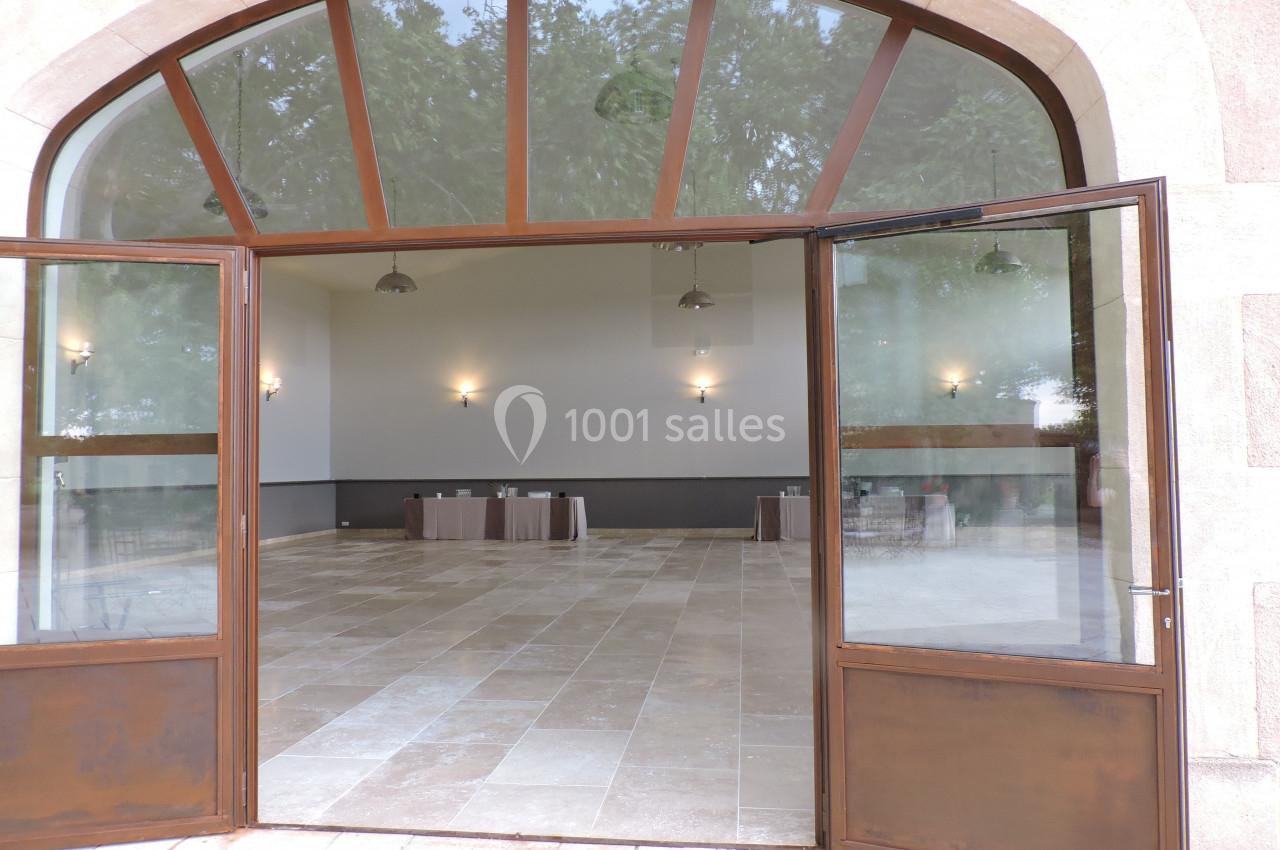 Salle vide avec sol en carrelage beige, murs blancs, tables couvertes en fond et grandes portes vitrées ouvertes.