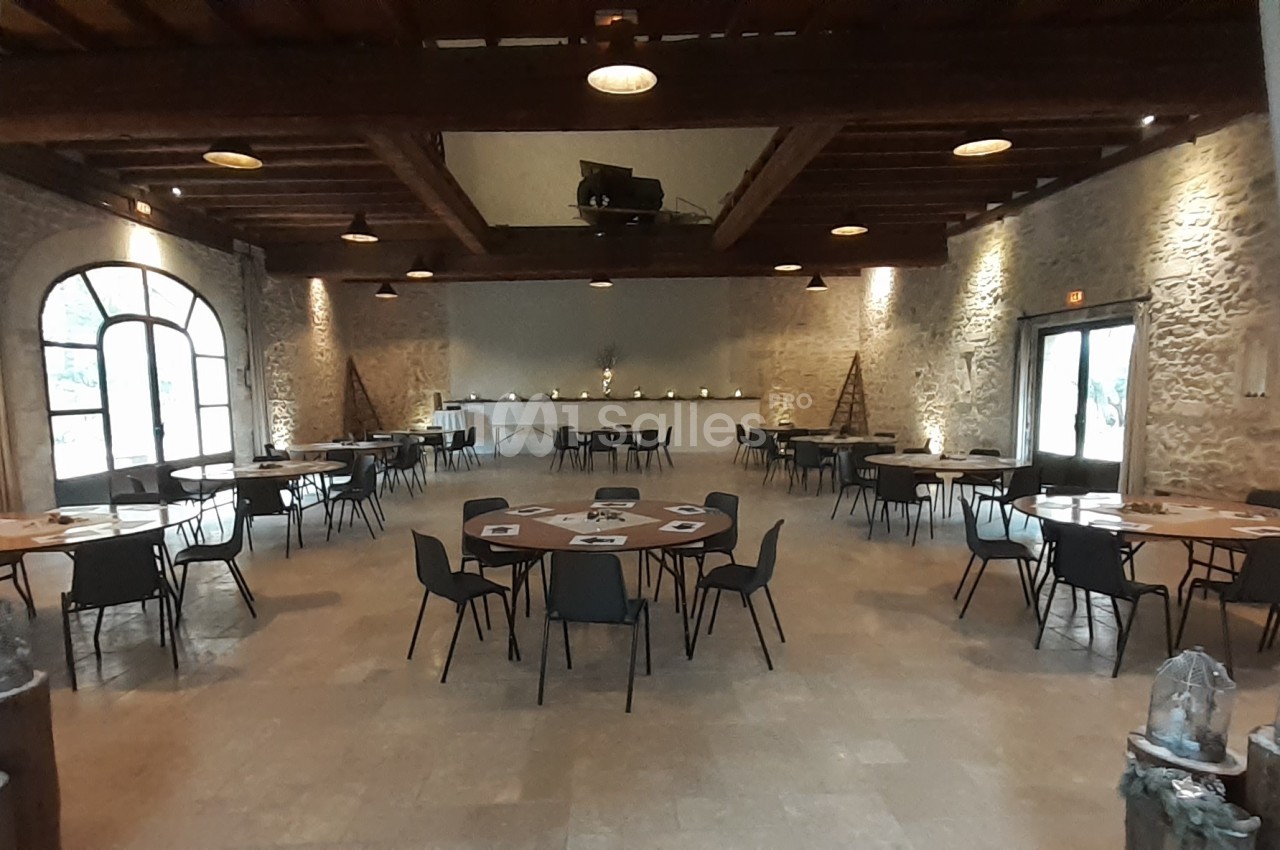 Salle spacieuse avec murs en pierre, tables rondes dressées et éclairage chaleureux, prête pour un événement.