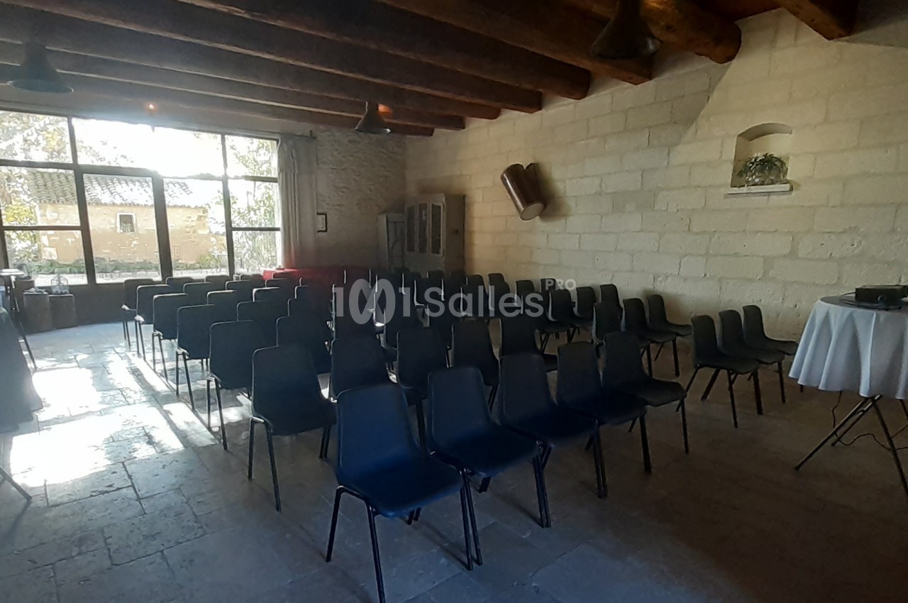 Salle avec des rangées de chaises noires vides, éclairée par une grande fenêtre donnant sur un extérieur arboré.