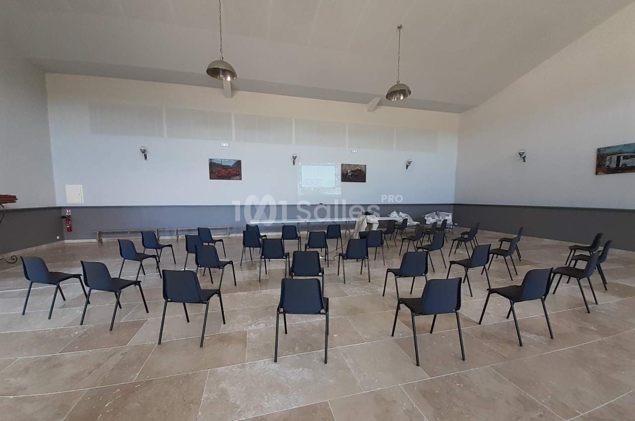 Salle vide avec des rangées de chaises noires disposées face à un écran de projection sur un mur clair.