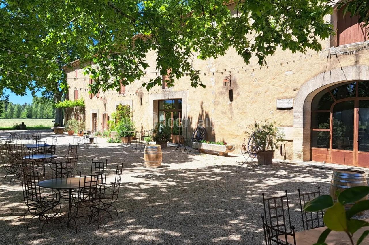 Cour ombragée d'une bâtisse en pierre avec tables et chaises en métal, entourée de végétation et baignée de lumière…