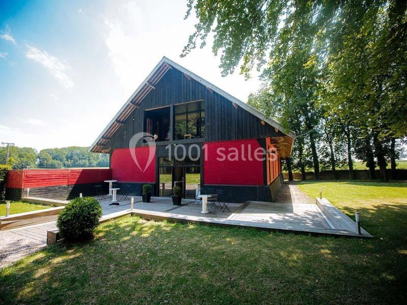 Bâtiment moderne en bois et rouge avec terrasse, entouré de pelouse et d'arbres sous un ciel dégagé.