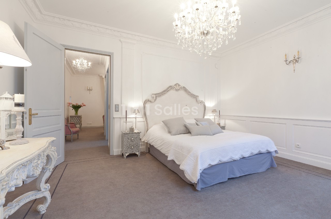 Chambre lumineuse avec lit double, murs blancs, lustre en cristal et mobilier classique dans un décor élégant.