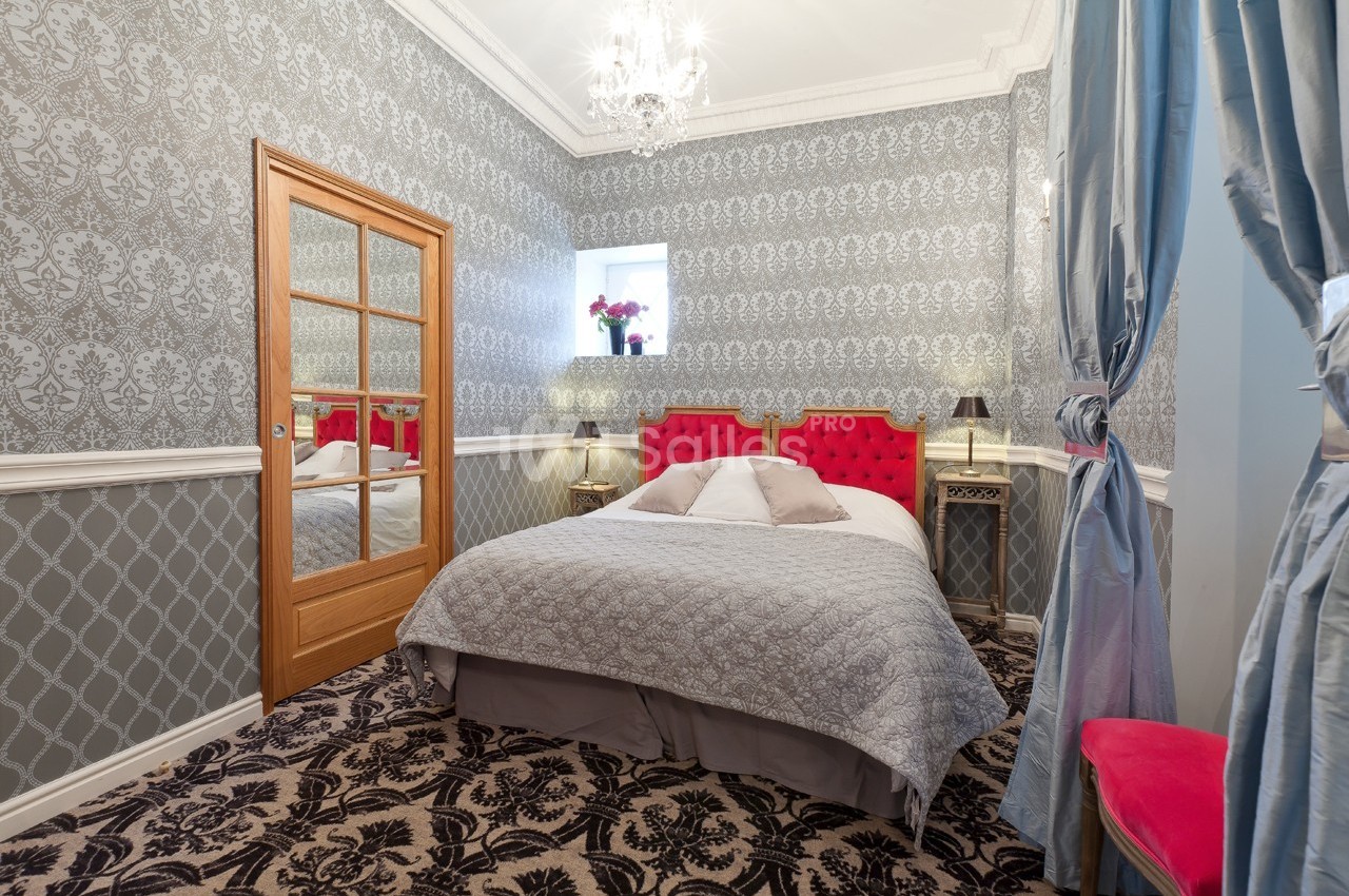 Chambre élégante avec lit double, tête de lit rouge, murs à motifs, lustre et rideaux bleus.