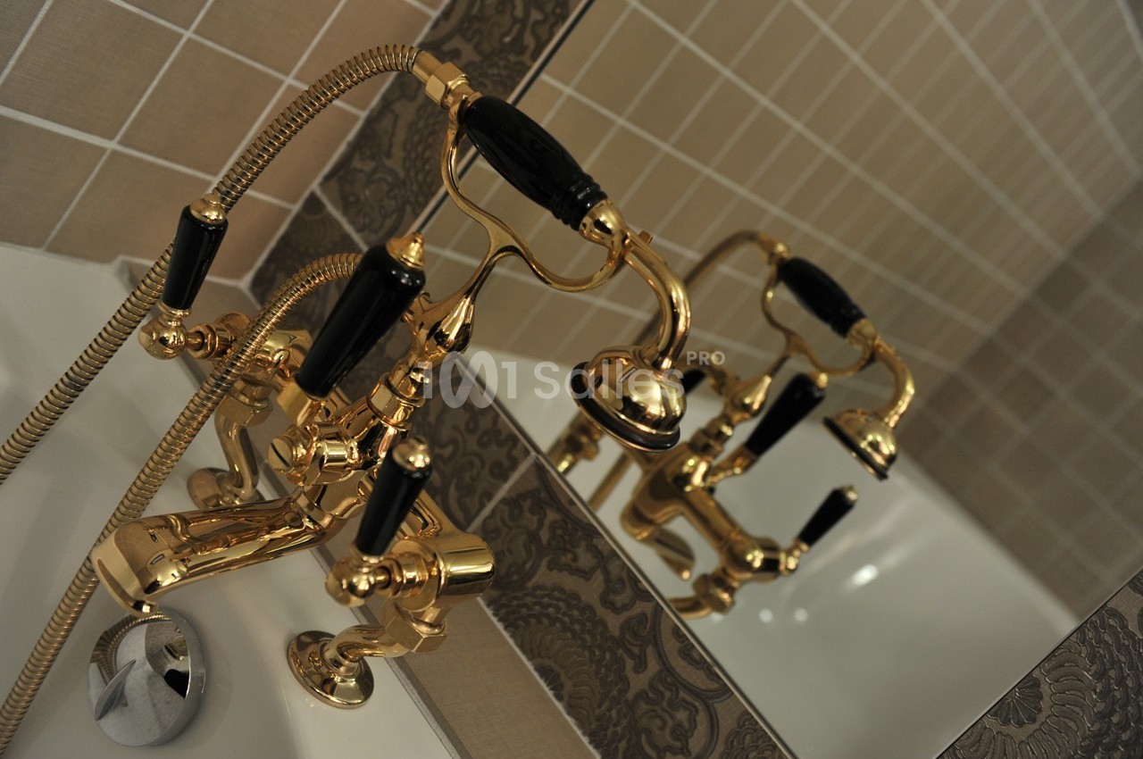 Robinetterie dorée avec poignées noires et douchette, installée sur le bord d'une baignoire devant un miroir.