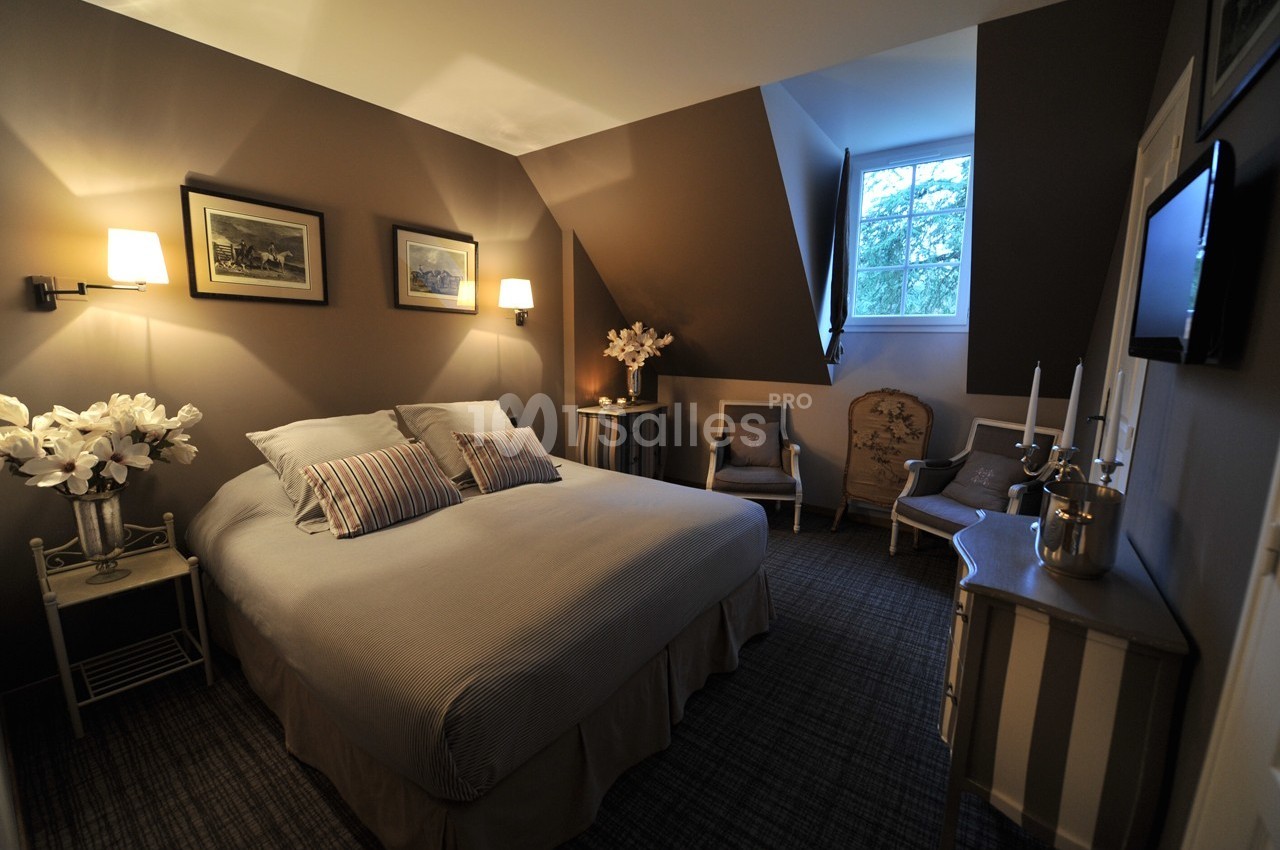 Chambre décorée avec des tons neutres, lit double, fauteuils, éclairage doux et fenêtre donnant sur des arbres.