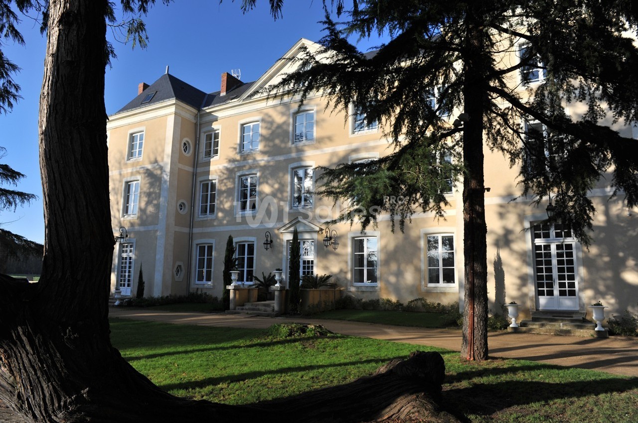 Façade d'un grand bâtiment beige à plusieurs étages, entouré d'arbres et d'une pelouse sous un ciel dégagé.
