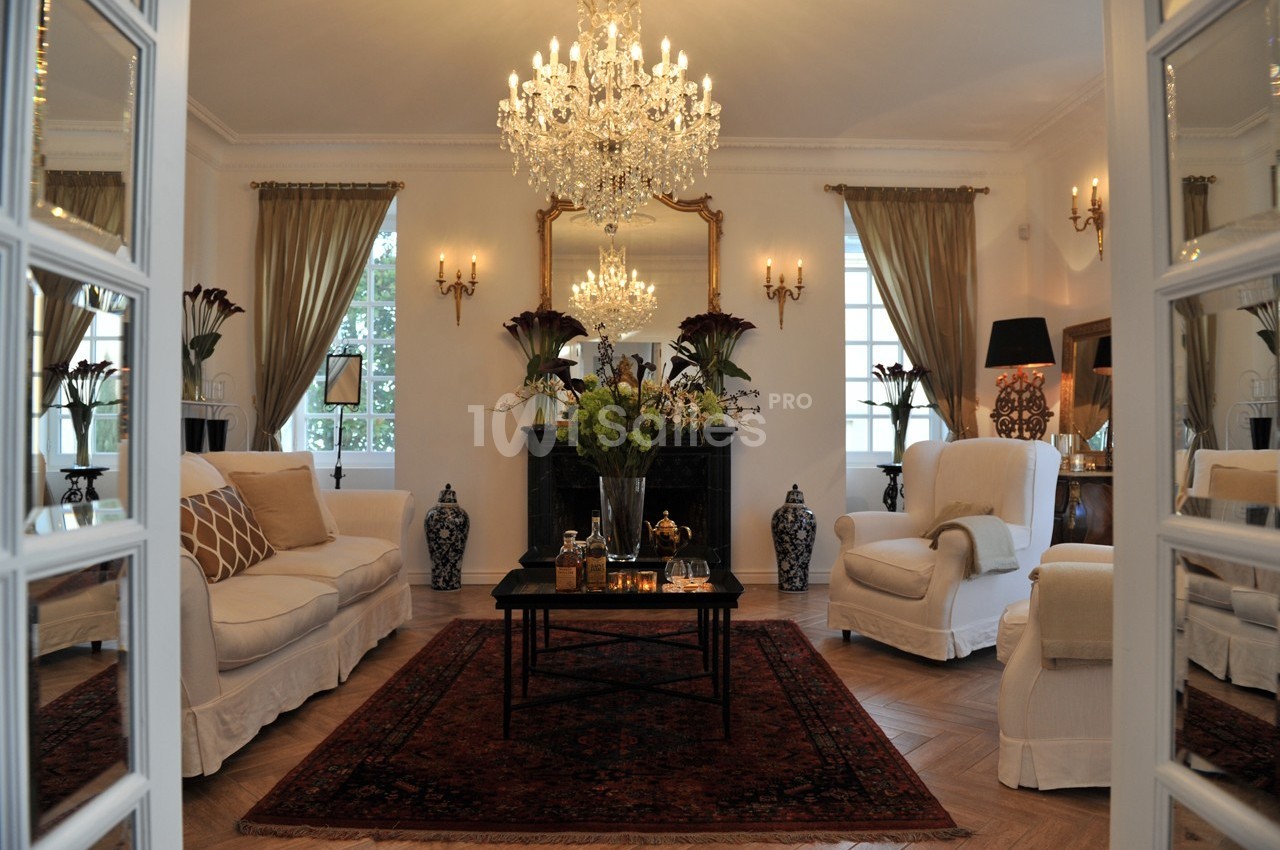 Salon élégant avec lustre en cristal, meubles classiques, tapis rouge et grandes fenêtres encadrées de rideaux.