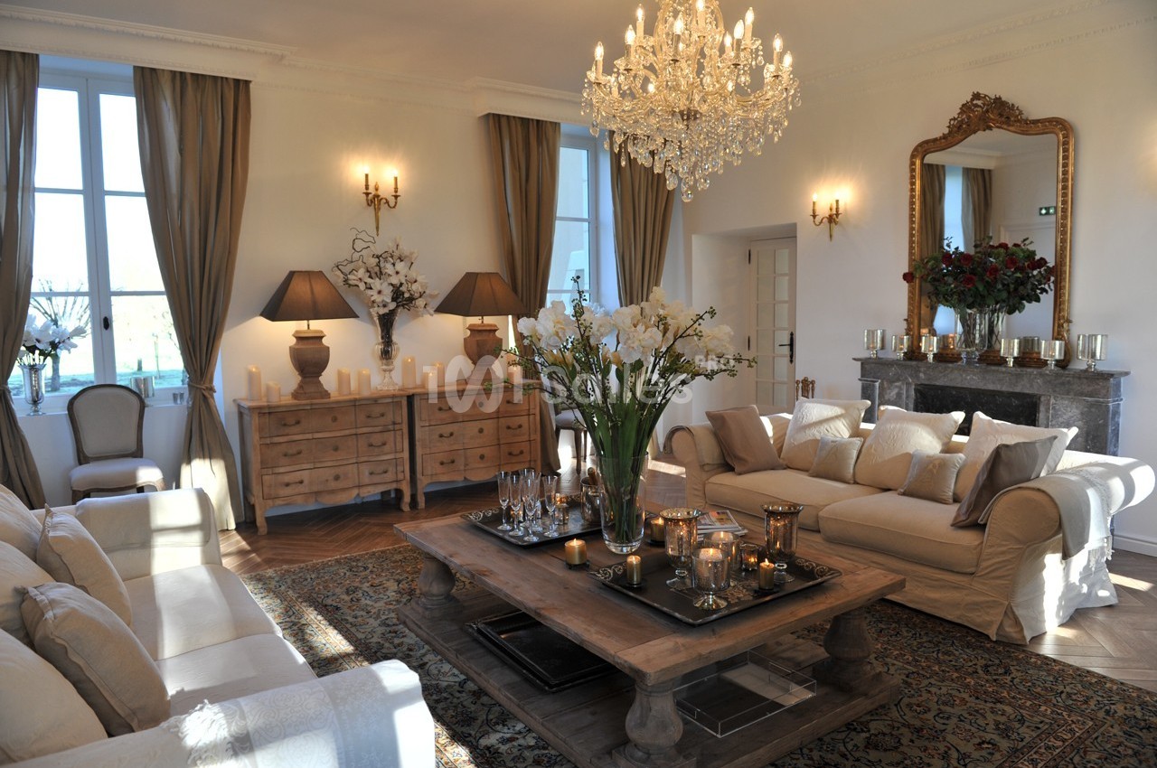 Salon élégant avec canapé beige, table basse en bois, lustre en cristal, cheminée et grandes fenêtres avec rideaux.