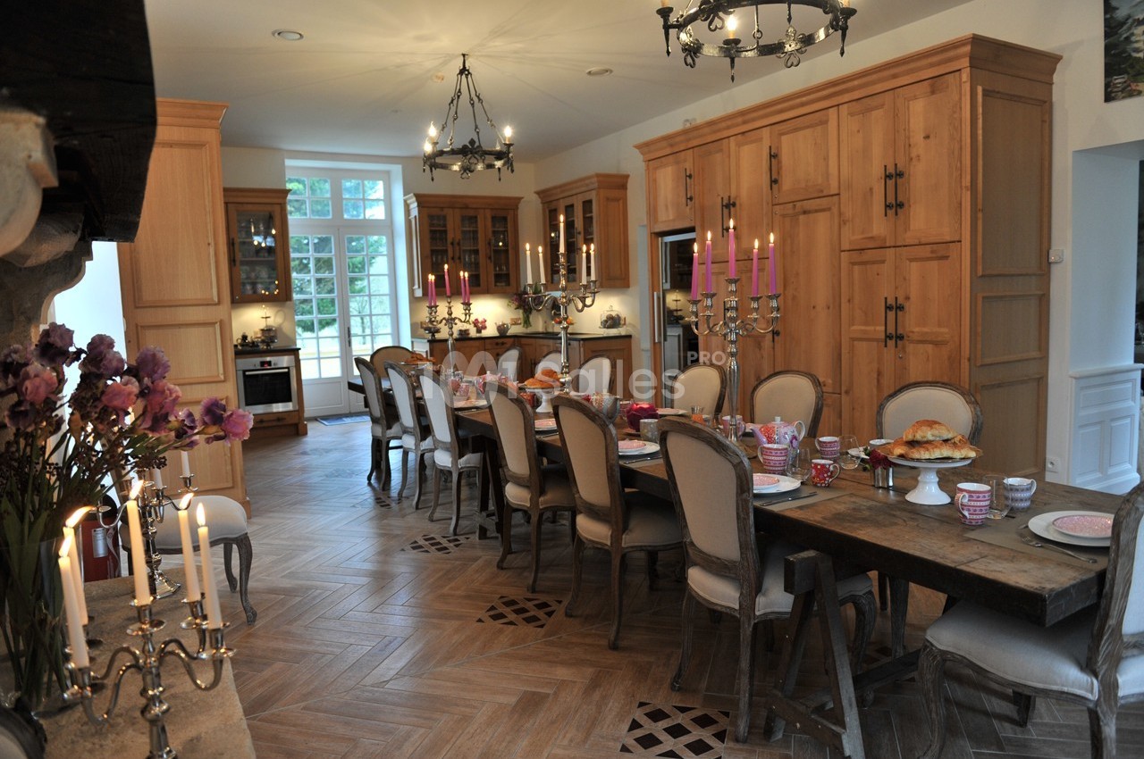 Grande cuisine lumineuse avec table en bois dressée pour un repas, entourée de chaises et décorée de chandeliers.