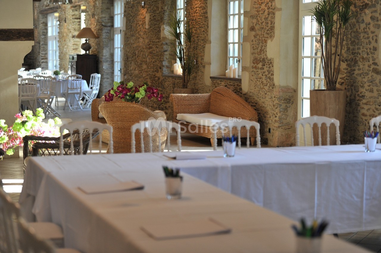 Salle lumineuse avec murs en pierre, tables blanches, chaises et décoration florale, ambiance conviviale et élégante.