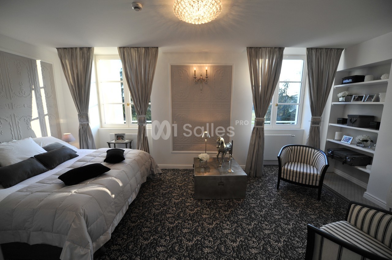 Chambre lumineuse avec lit double, fauteuils, tapis à motifs, rideaux gris et étagères blanches intégrées.