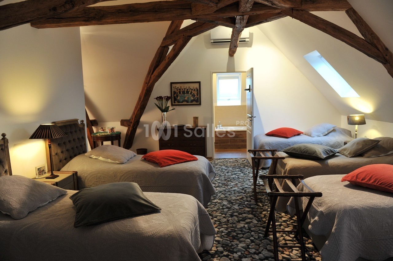 Chambre mansardée avec poutres apparentes, quatre lits simples, lumière naturelle et salle de bain attenante.