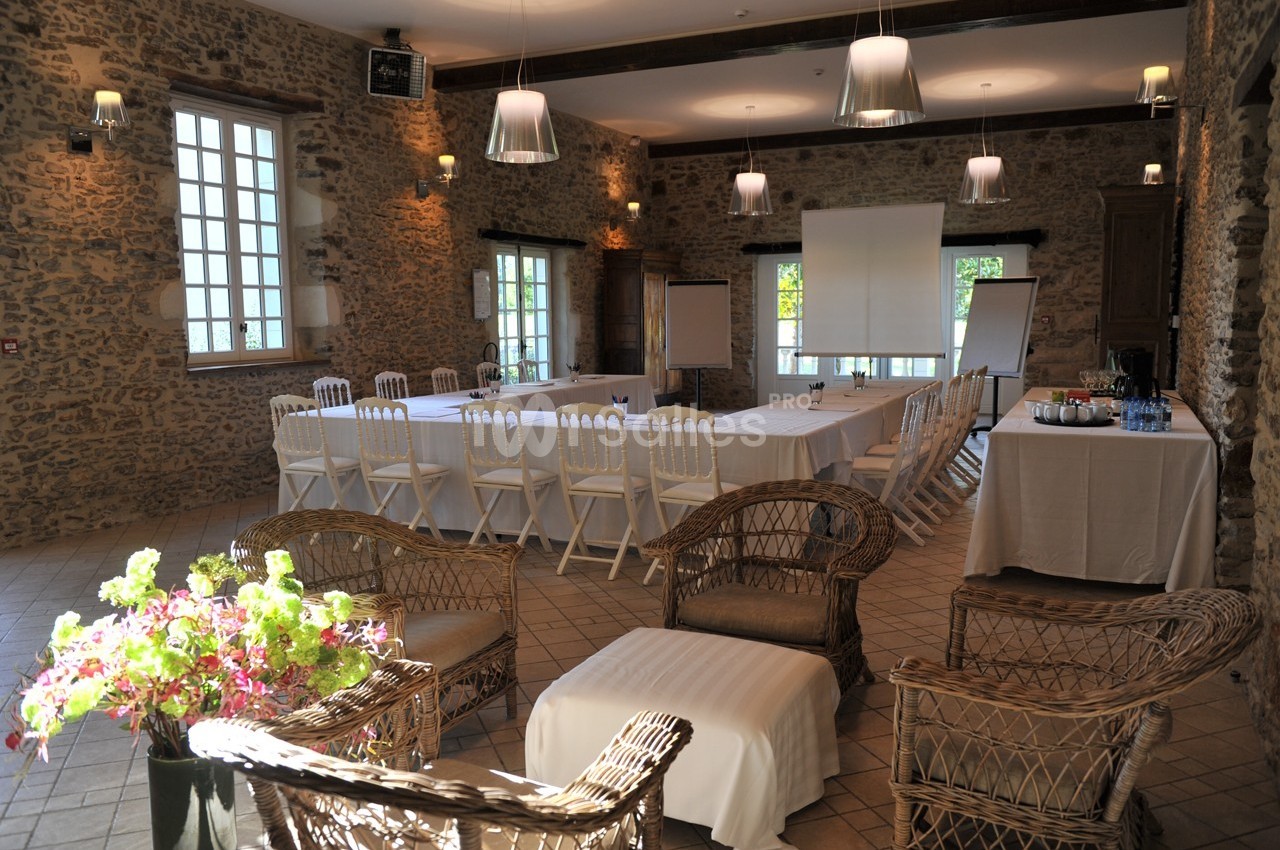 Salle de réunion lumineuse avec murs en pierre, tables disposées en U, chaises blanches et matériel de présentation.