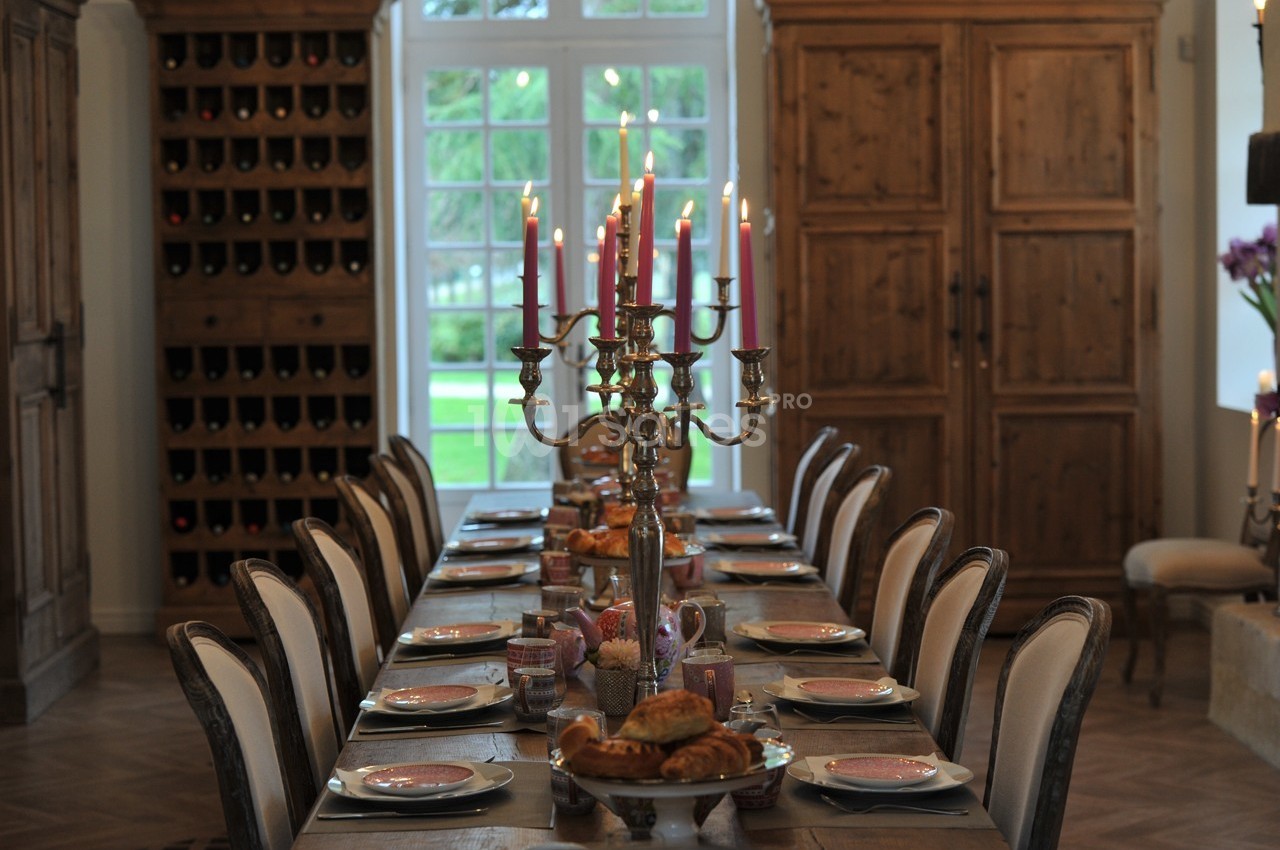 Grande table dressée avec des chandeliers allumés, vaisselle élégante et viennoiseries, dans une salle à manger rustique.