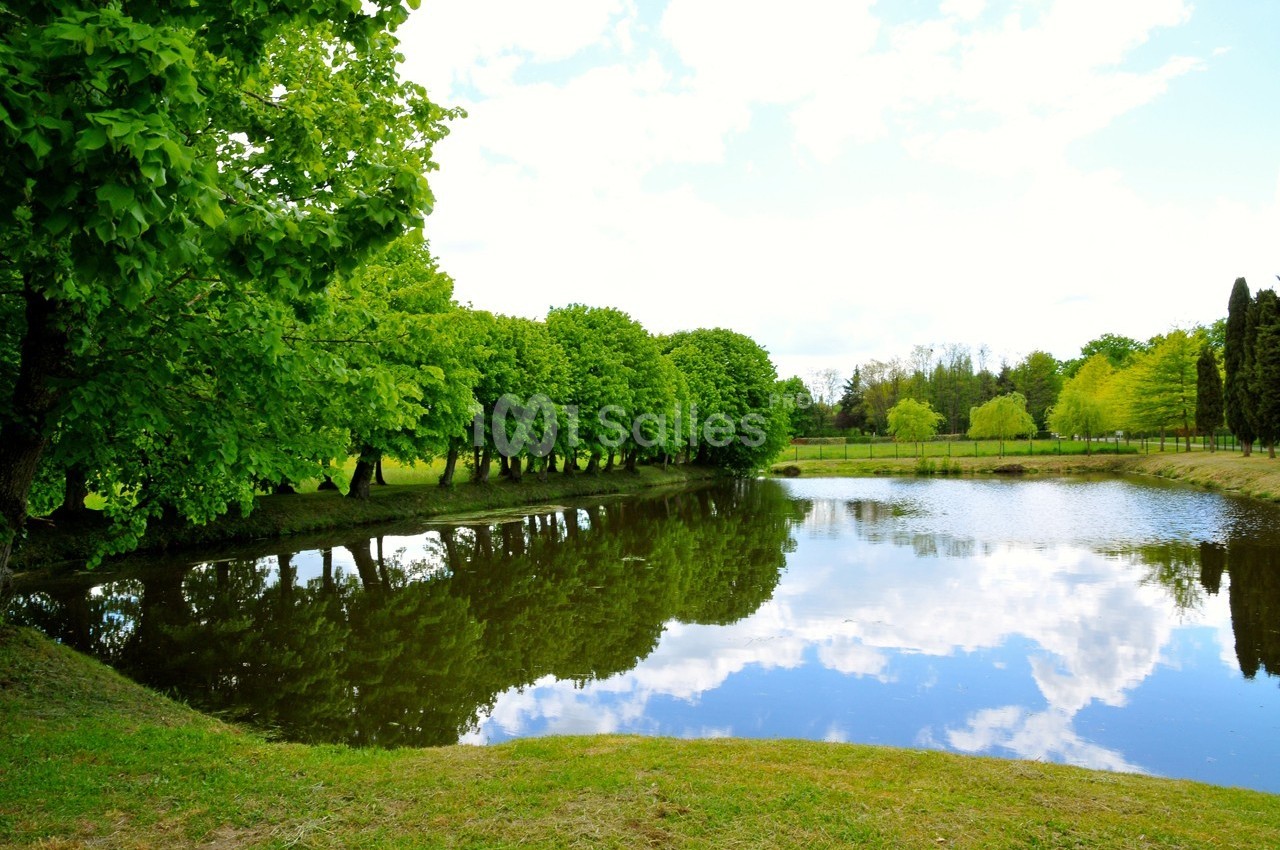 Étang entouré d'arbres avec leur reflet visible dans l'eau sous un ciel partiellement nuageux.