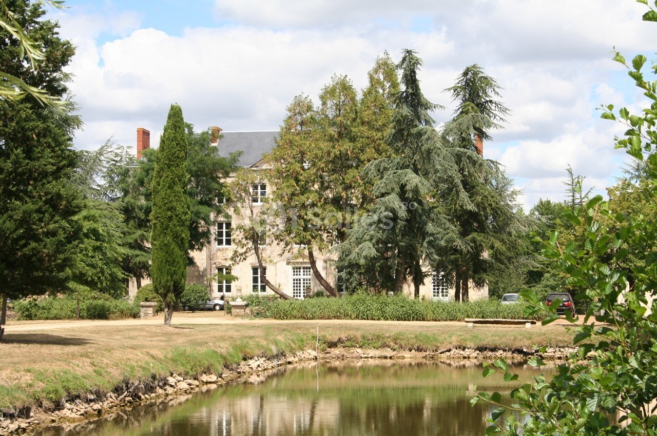 Façade d'une maison en pierre entourée d'arbres, avec un étang au premier plan sous un ciel partiellement nuageux.