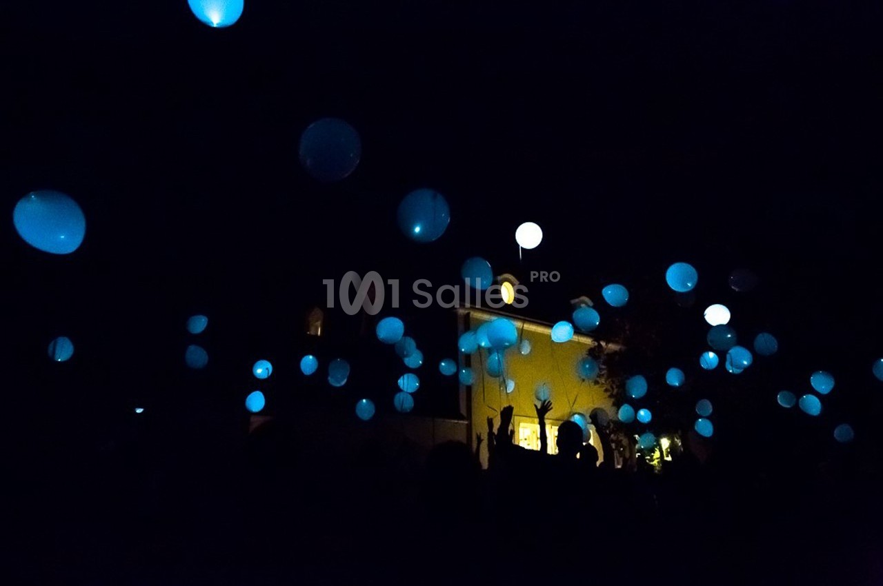 Des ballons lumineux bleus s'élèvent dans le ciel nocturne, entourés de silhouettes sombres et d'un bâtiment éclairé.