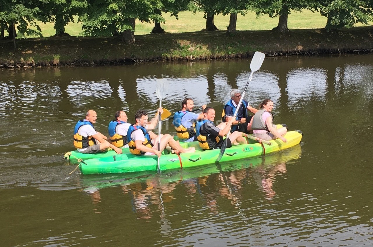 Des personnes pagayant sur un grand canoë vert et jaune sur une rivière entourée d'arbres.