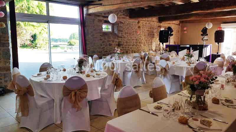 Salle de réception décorée pour un événement, avec des tables rondes dressées et des chaises ornées de nœuds en tissu beige.