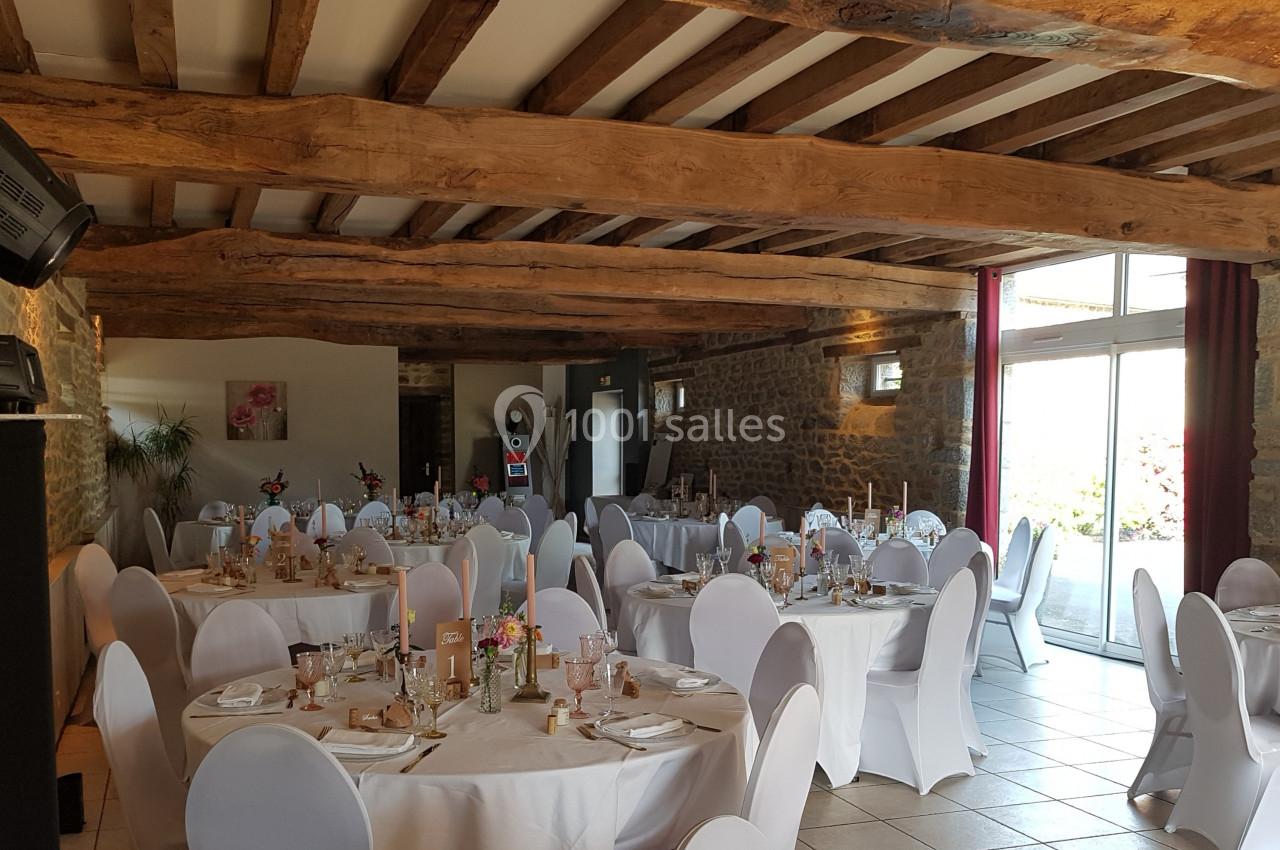 Salle de réception avec des tables rondes dressées, chaises blanches et poutres apparentes au plafond.
