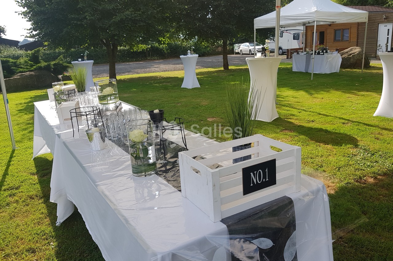 Table décorée en extérieur sous une tente, avec des éléments en verre, des plantes et une caisse en bois blanche.