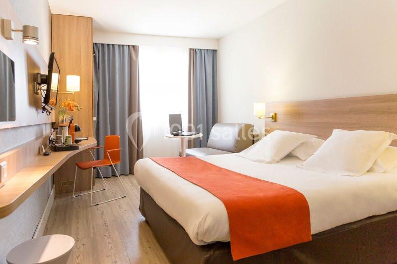 Chambre d'hôtel lumineuse avec lit double, bureau, chaise orange, canapé et grande fenêtre avec rideaux.
